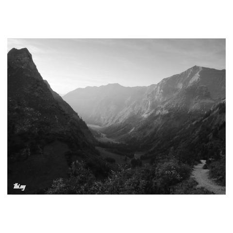 B&W photo of a beautiful curved alpine valley, seen from slightly higher up and just before sunset. A bright light shaft is illuminating the steep mountain slopes on the right in the background. Most of the foreground is in shade already. A small foot path is leading down through bushy vegetation. (Much) further in the distance we can see it winding through the forest and meadows on the valley floor towards the light. Hazy sky.