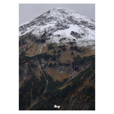 Portrait photo of a large mountain with the rocky upper third dusted in fresh snow and the steep slopes below a mixture of cliffs, meadows and forest, in beautiful rich autumn colors. Plain gray sky.