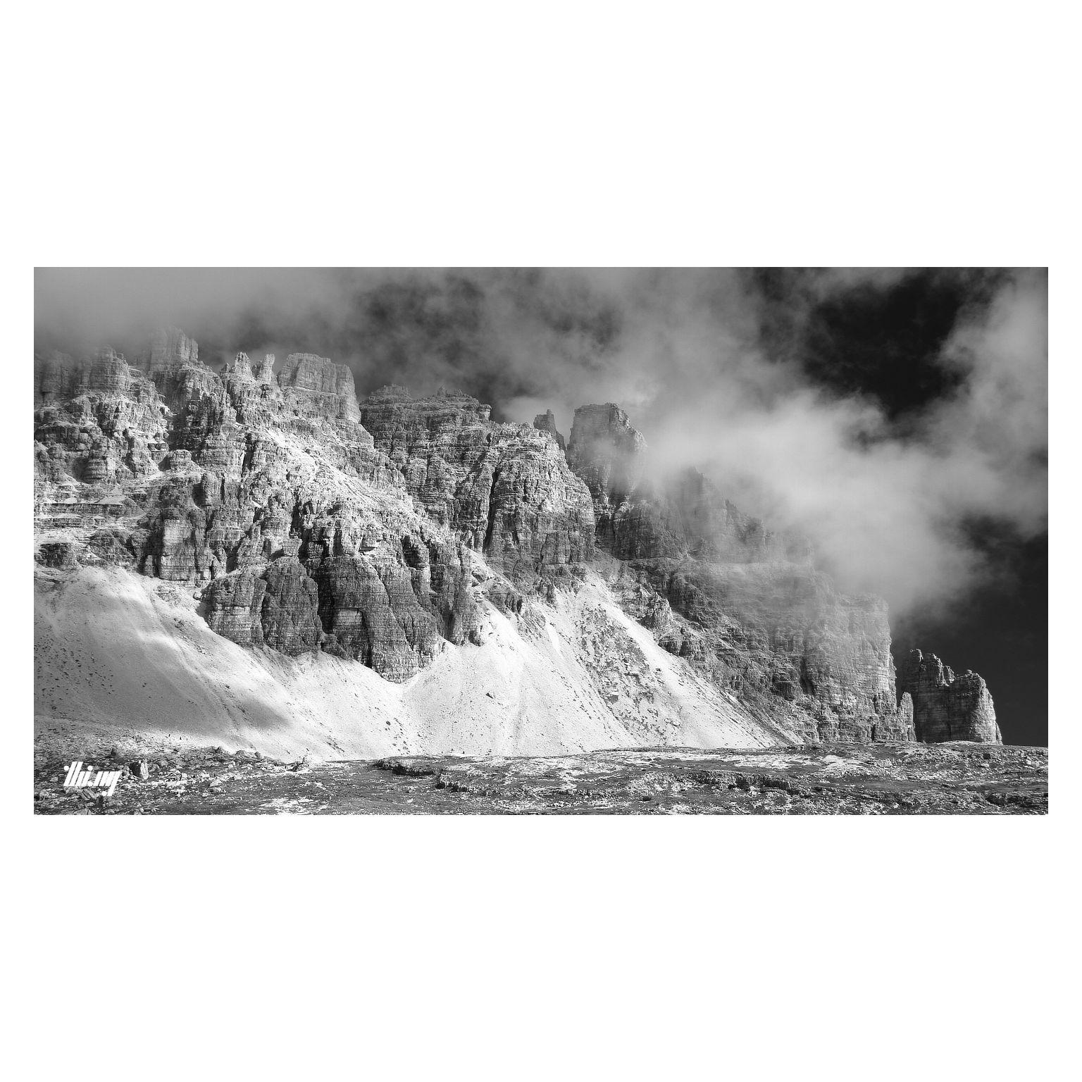 Wide-format B&W photo of a mountain group in the Dolomites with characteristic rock tower & needle structures and large/steep talus fields further below. A small rocky plateau is in the foreground, hiding a steep drop off at mid distance. Clouds are forming around the peaks and are drifting upwards against a dark sky. The cloud shadows are helping to create extra drama.